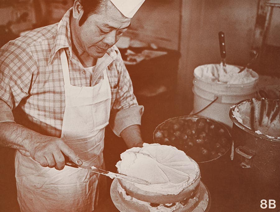 A black-and-white photograph of a baker adding frosting to a cake.