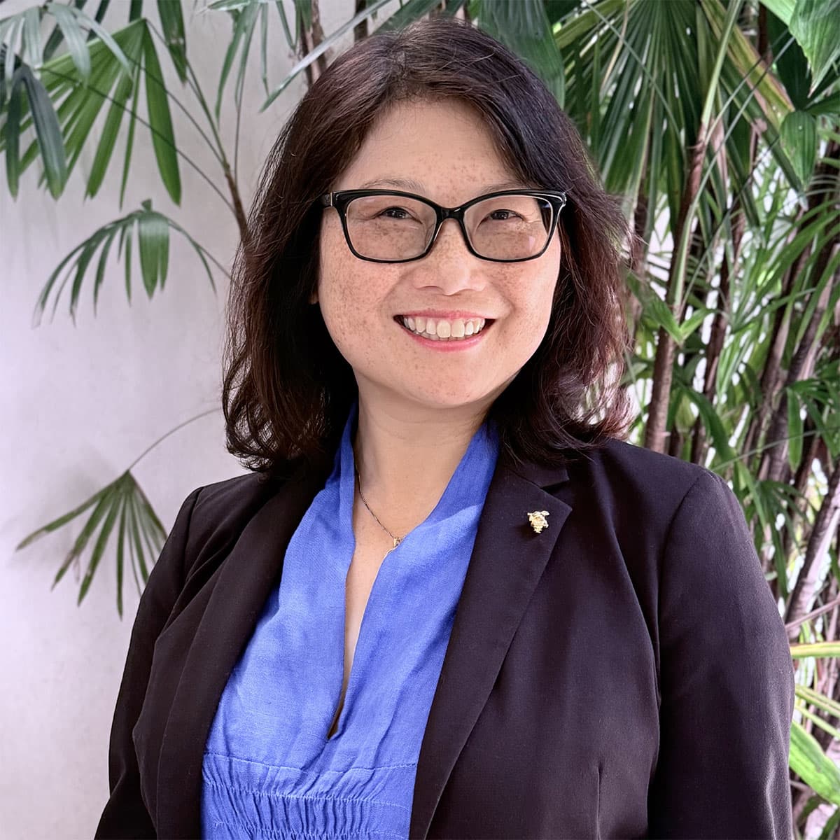 A person in a blue top and dark blue jacket smiles for the camera in front of a tropical plant.