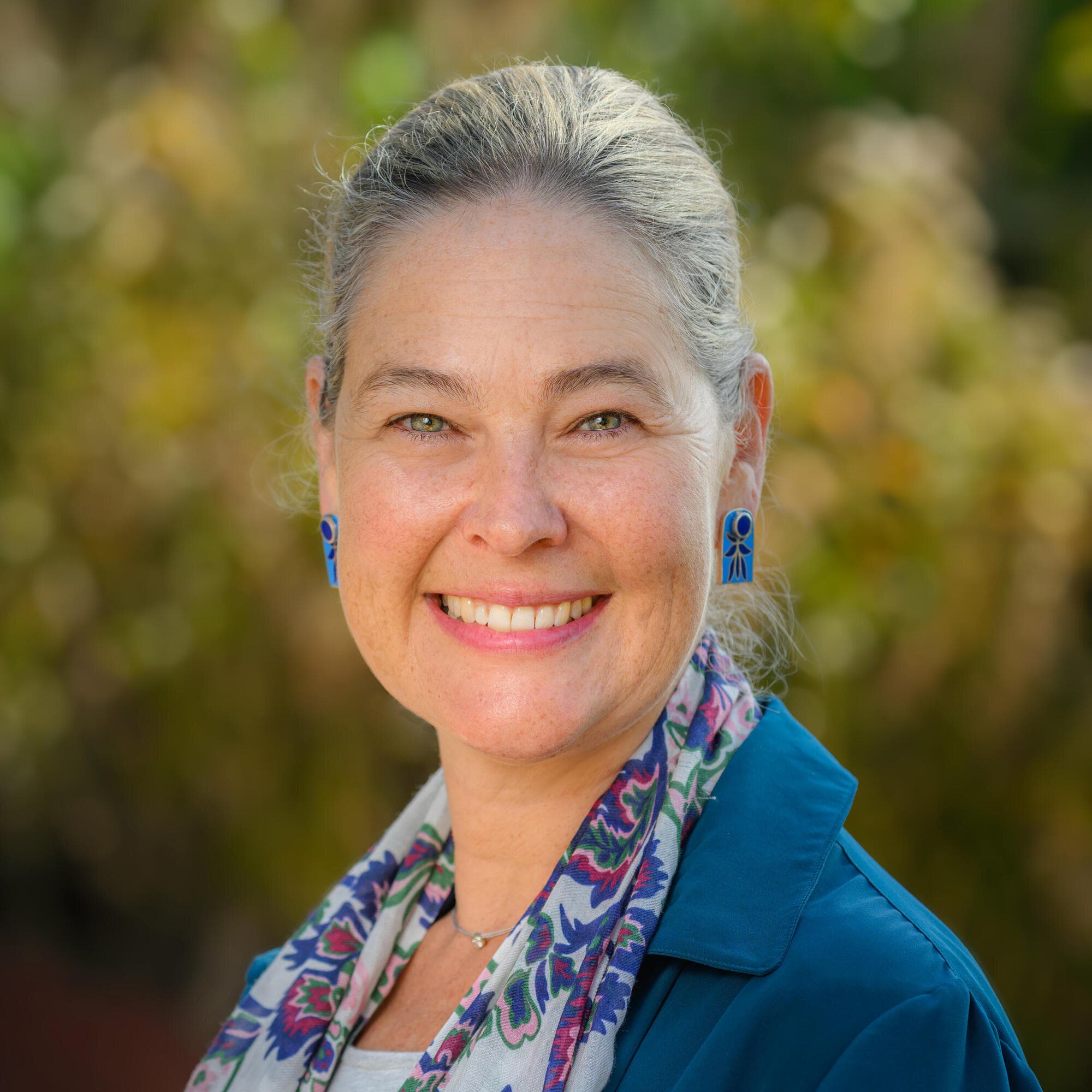 Person in a blue blazer and patterned scarf smiling outdoors.