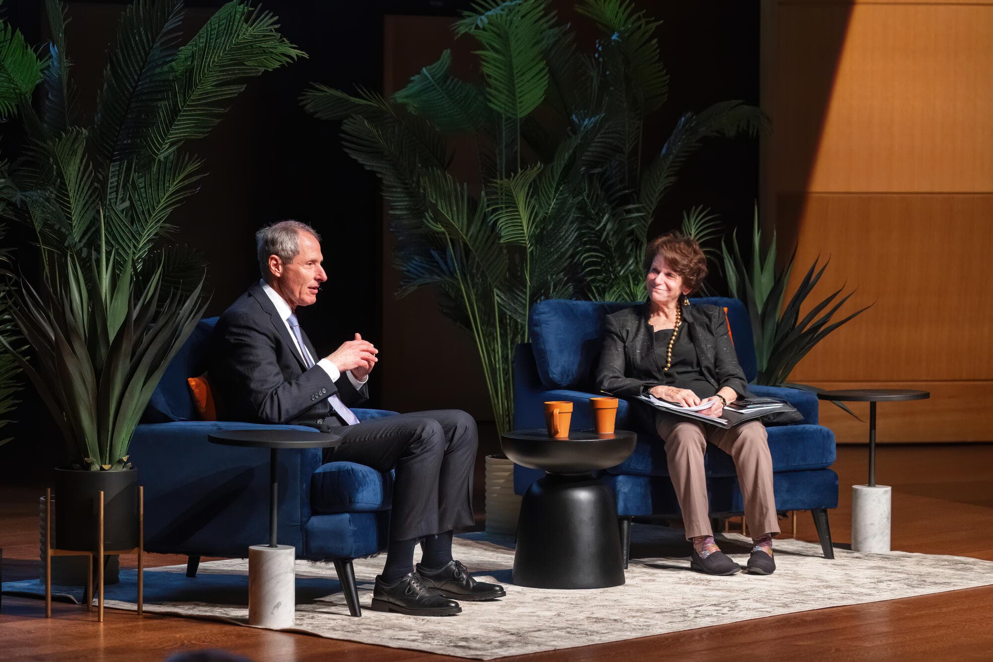 Two people sit on deep blue fabric chairs on a stage. One listens while the other speaks.