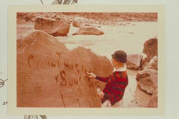 Dock Marston marks for the photographers the inscription made by Harry McDonald of the Best Expedition after they locked a boat on the flat rock at upper left on July 21, 1891.  Rapid No. 16 at Mile 204 1/2