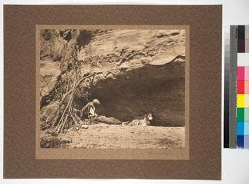 Old Navajo man taking shelter against a sandstone wall, with a dog at his feet