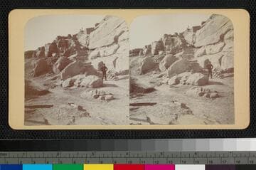 A Pueblo Indian stands near stairs leading to an unidentified pueblo on top of a mesa