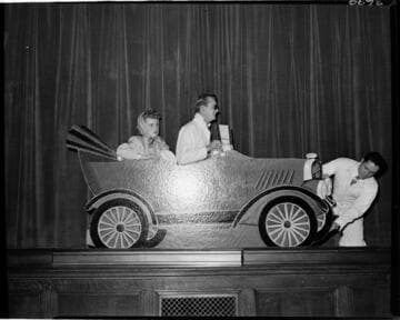 Performance of skit at Edison auditorium.  Man and woman in cardboard car with another man fixing a flat on the car