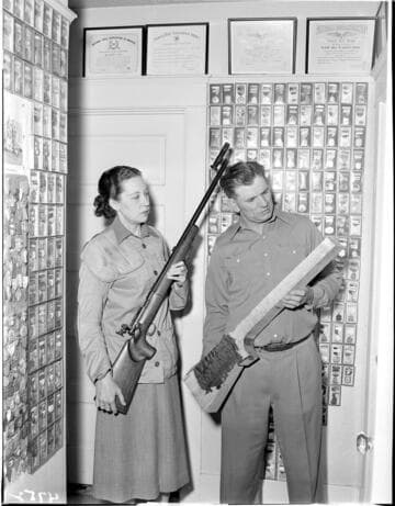 Man and woman in room filled with framed awards and medals for marksmanship.  Woman is holding a sharpshooters rifle and man is holding a blank un