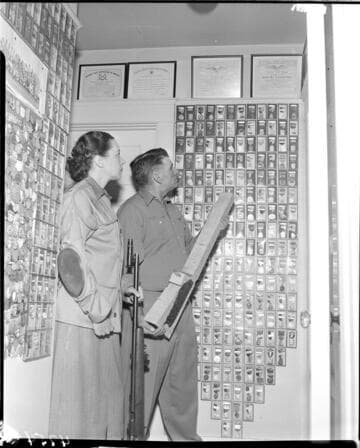 Man and woman in room filled with framed awards and medals for marksmanship.  Woman is holding a sharpshooters rifle and man is holding a blank un