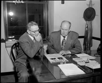 Two men at a desk looking over FBI wanted poster
